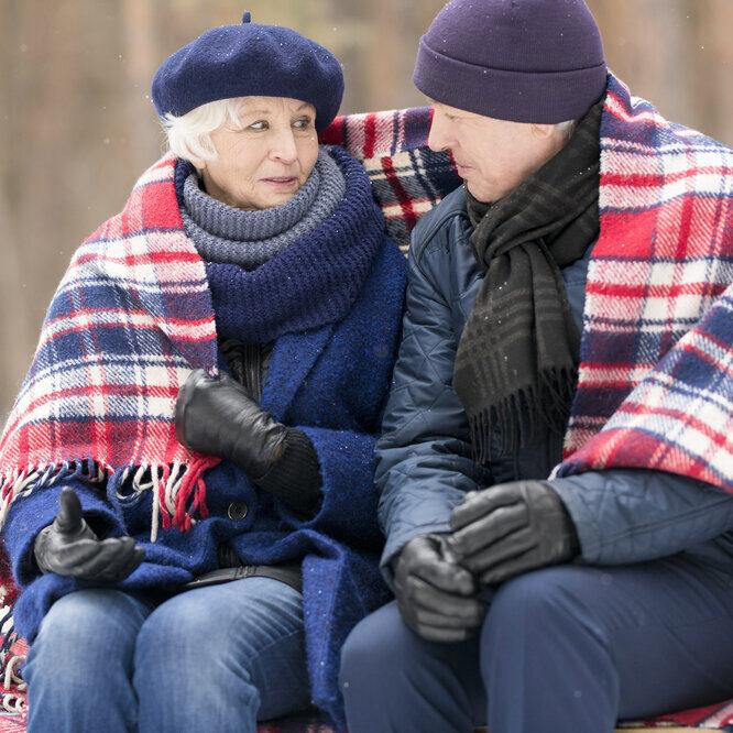 Portrait of loving senior couple sitting on bench in winter sharing blanket and chatting, copy space